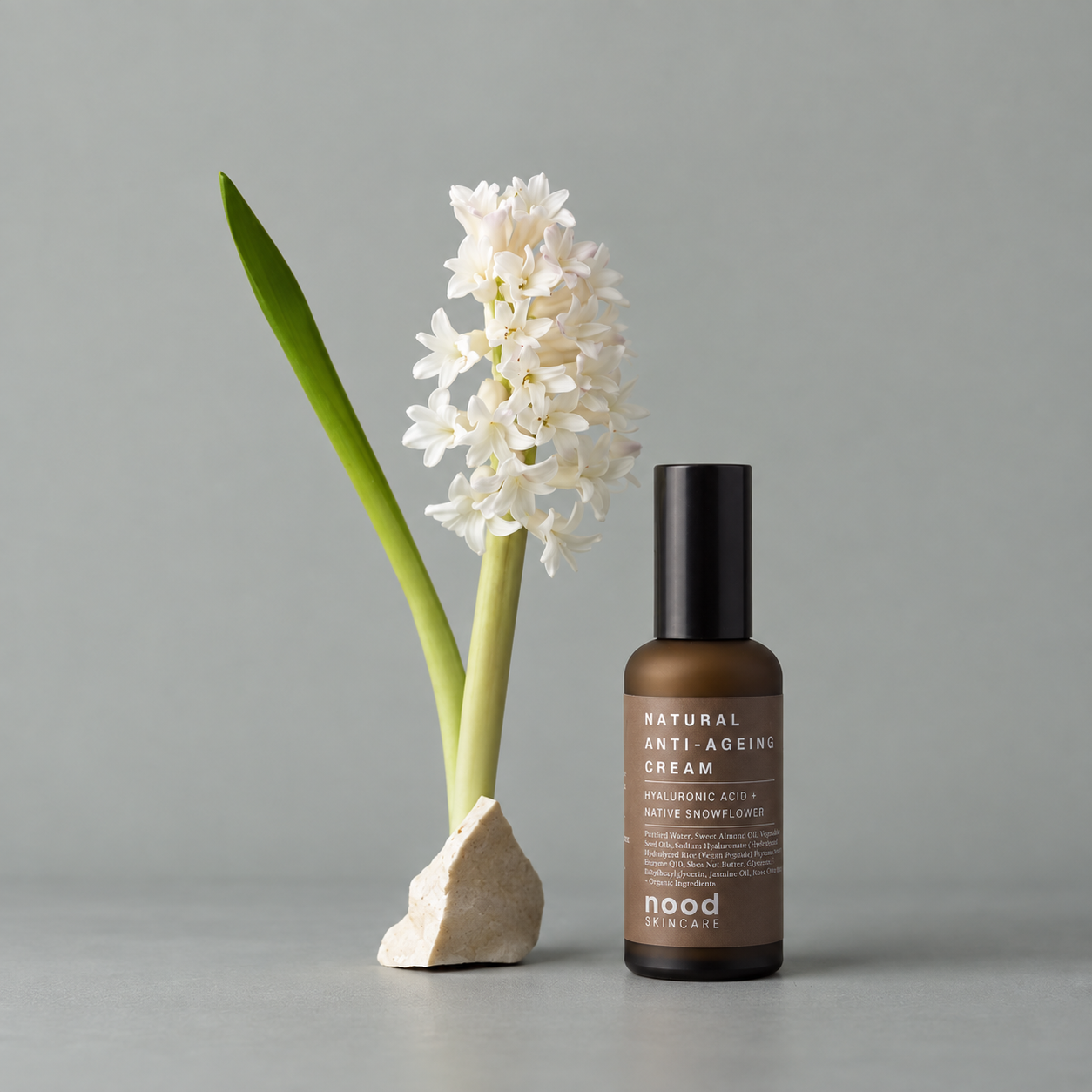 A bottle of Nood mood Natural Anti-Ageing Cream stands next to a white hyacinth flower on a light stone base against a solid gray background.