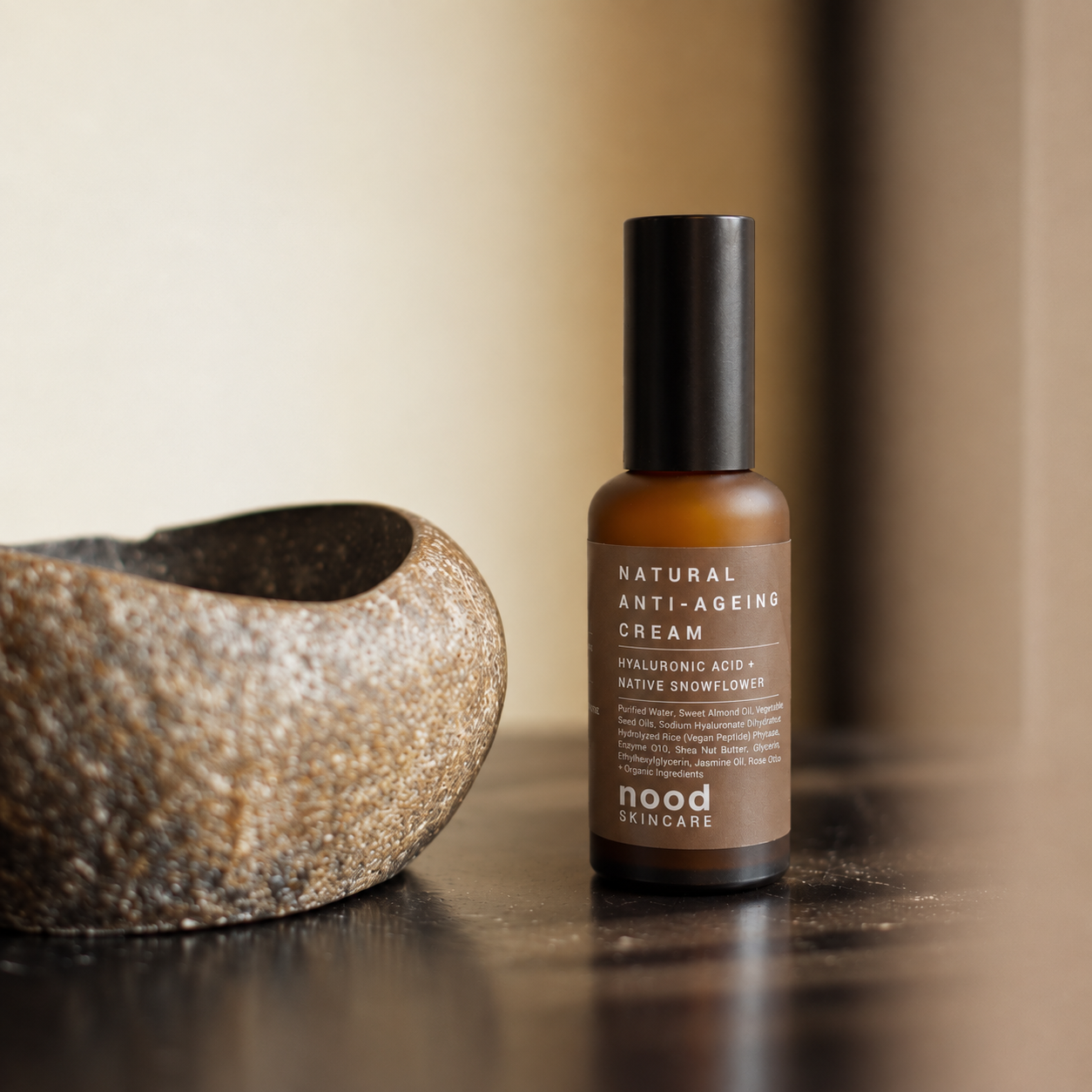A bottle of Nood Skincare Natural Anti-Ageing Cream stands on a dark wooden surface next to a textured stone bowl, with a warm beige background.