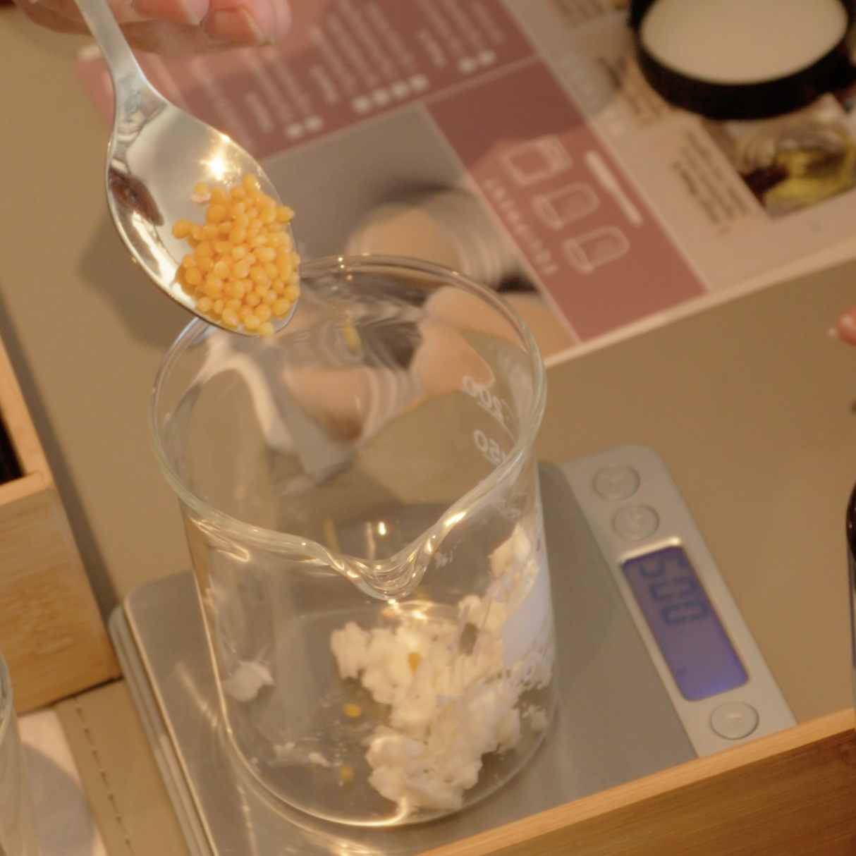 Person adding small yellow granules to a glass container on a kitchen scale.